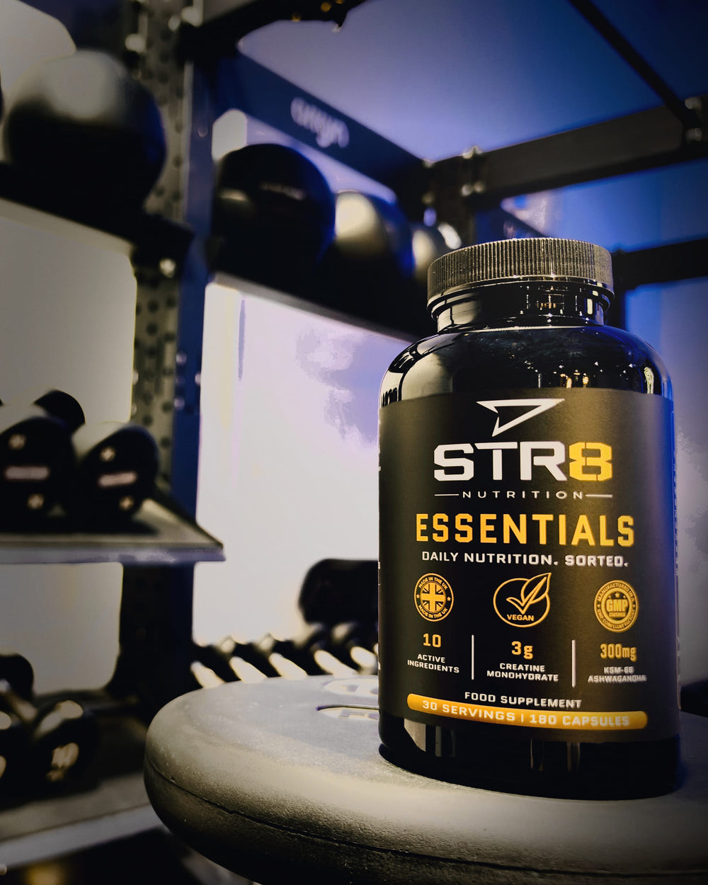 Essentials bottle on a weight plate beside a rack of dumbbells