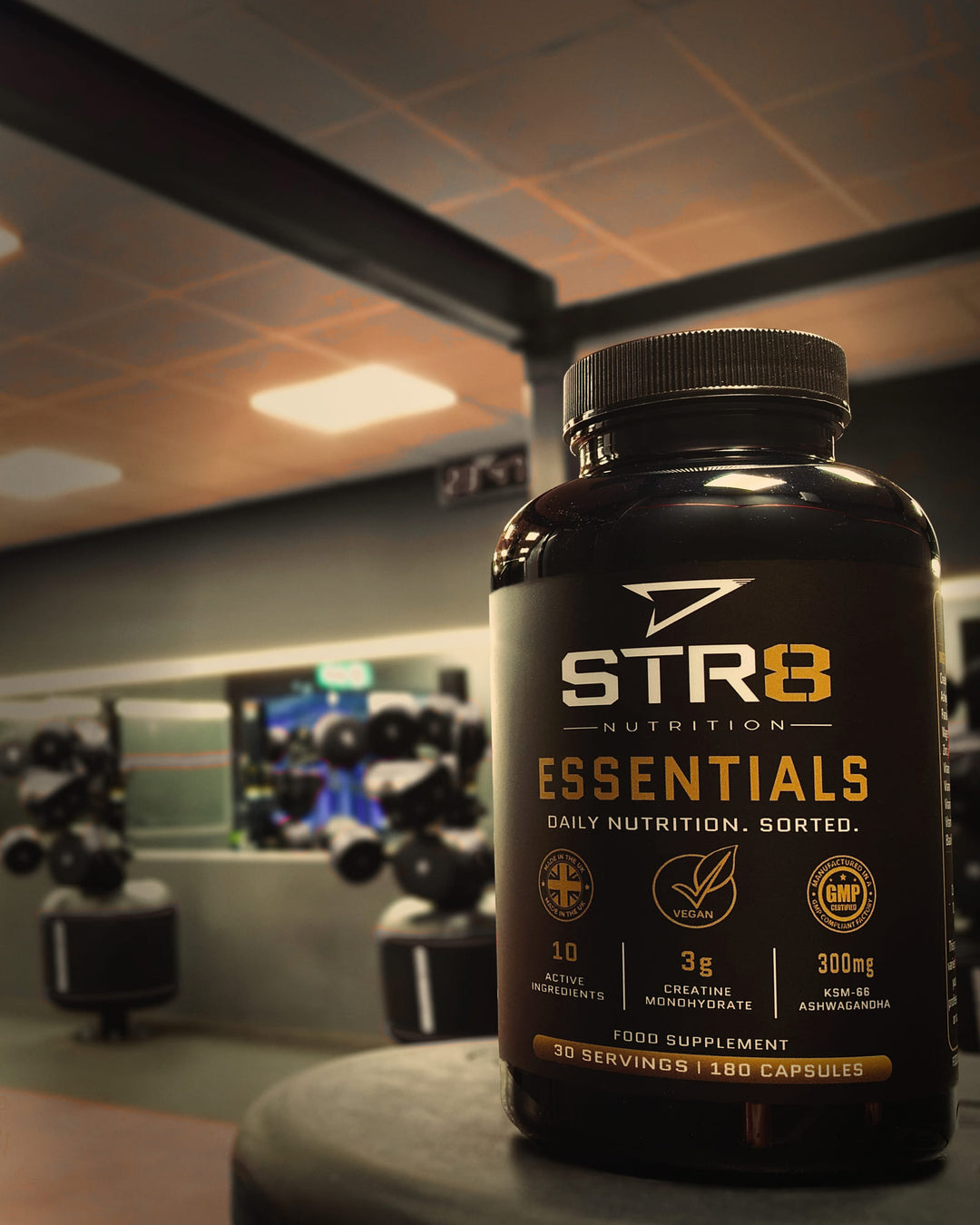Essentials bottle on a bench in a dark gym, punchbag and dumbbell racks behind